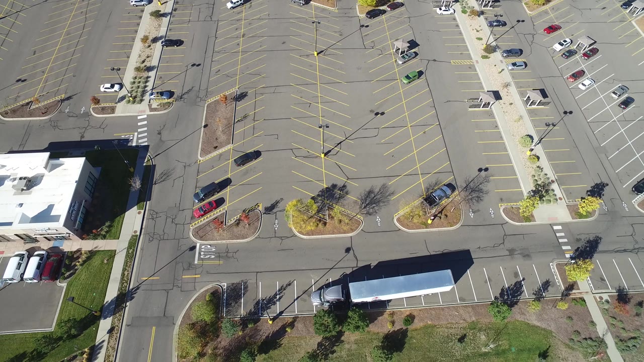 Second overhead frame in Hidden Lake, Colorado with the parking lot centered higher in view, the white-roofed building lower left, parked cars along the curb, and no visible people.