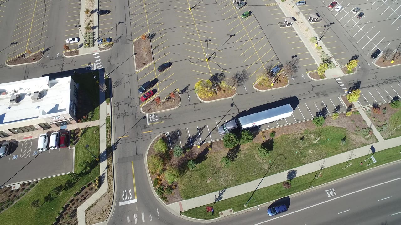 High aerial frame over Hidden Lake, Colorado showing a street corner, landscaped grass strip, parking lot rows, low buildings, parked cars, and no visible people.