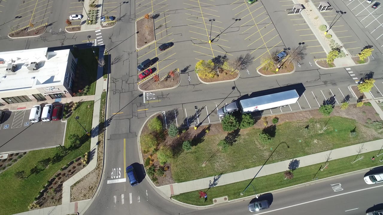 Drone-style overhead frame in Hidden Lake, Colorado with the long white trailer centered near a grass strip, planted islands, parking stalls, a road edge, and no visible people.