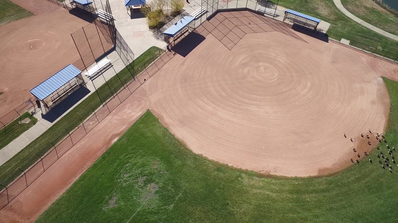Aerial view over an empty baseball field in Hidden Lake, Colorado, with the sandy infield high in frame, green grass below, blue-roofed shelters, fencing, and no visible people.