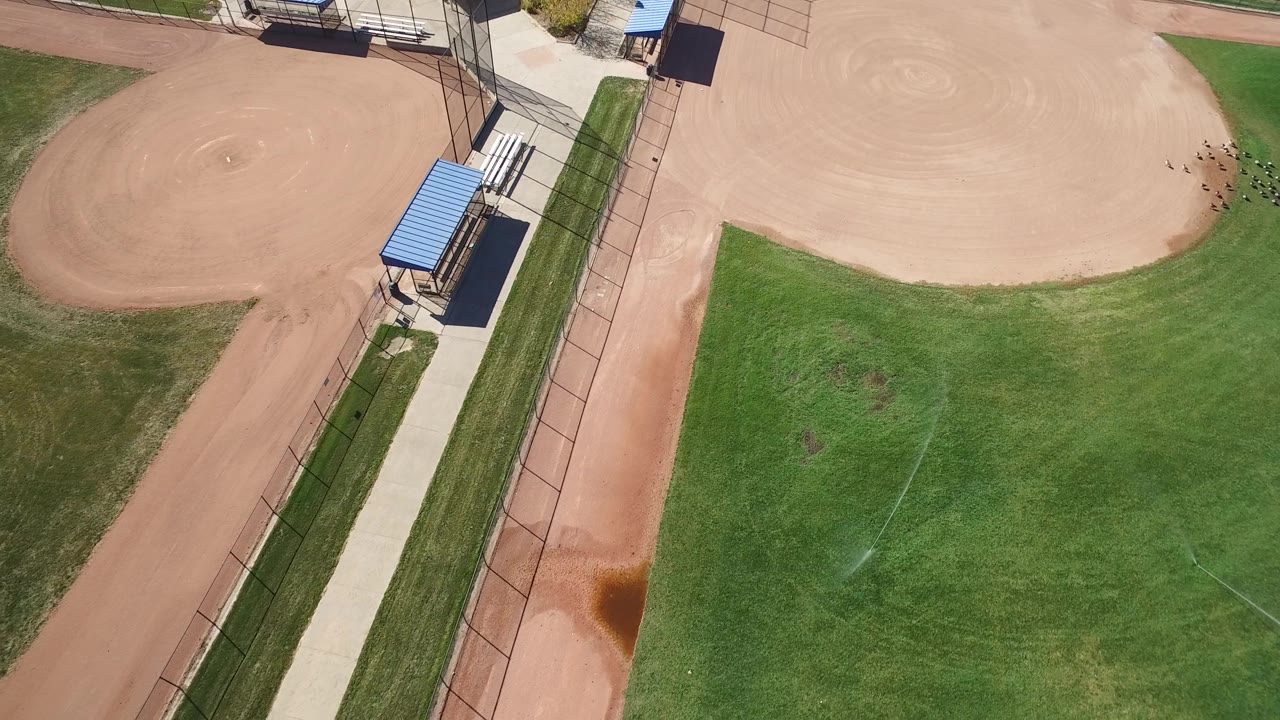 Aerial frame in Hidden Lake, Colorado with the left baseball diamond partly cropped, the upper infield visible, a narrow concrete path, fencing, blue dugout roofs, and small distant people near the top right.