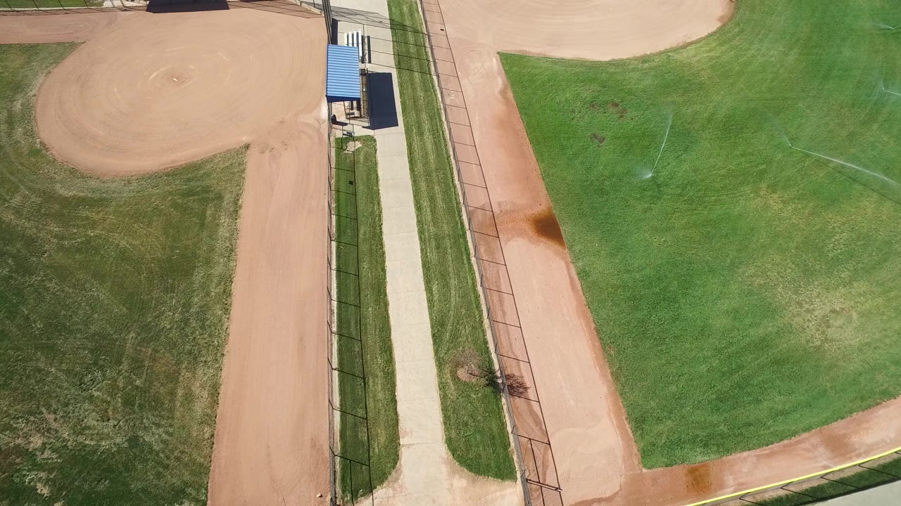 Top-down view in Hidden Lake, Colorado showing a tan baseball infield, green grass, a straight paved walkway, fencing, a blue-roof shelter, and no visible people.
