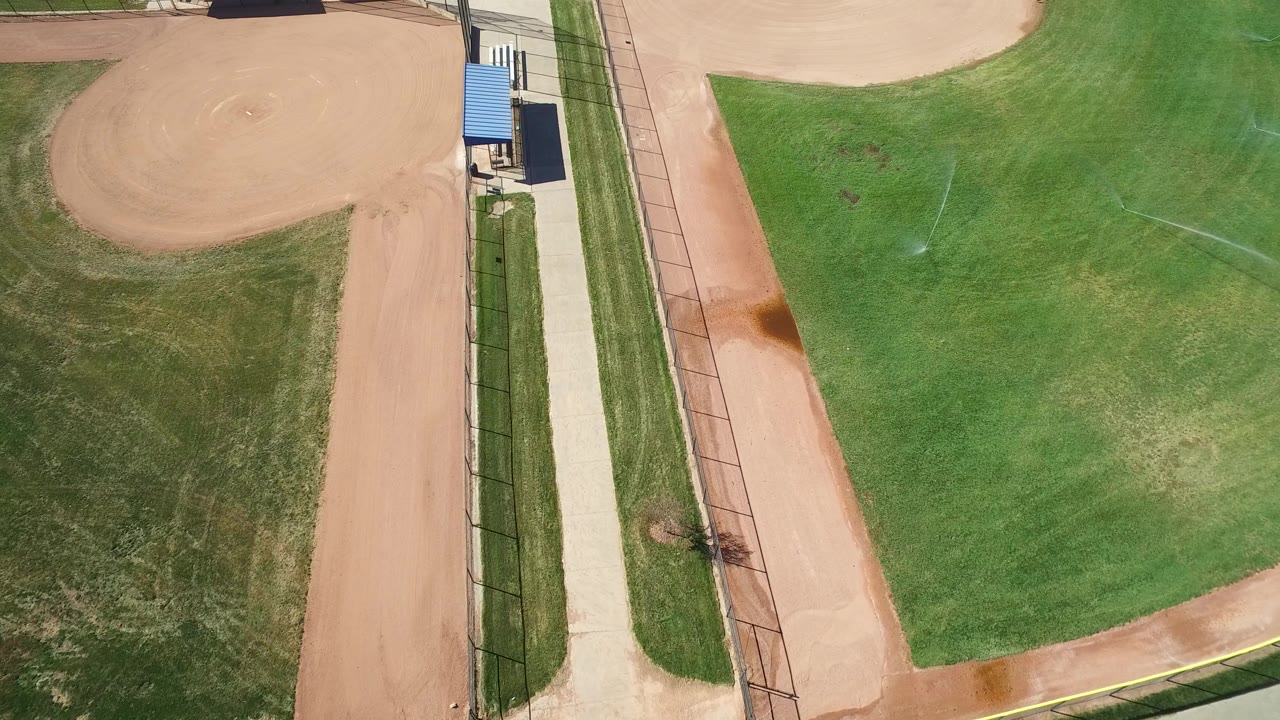 Aerial frame over Hidden Lake, Colorado with the infield at upper left, a central grass area, sprinkler arcs, a paved path, fencing, and no visible people.