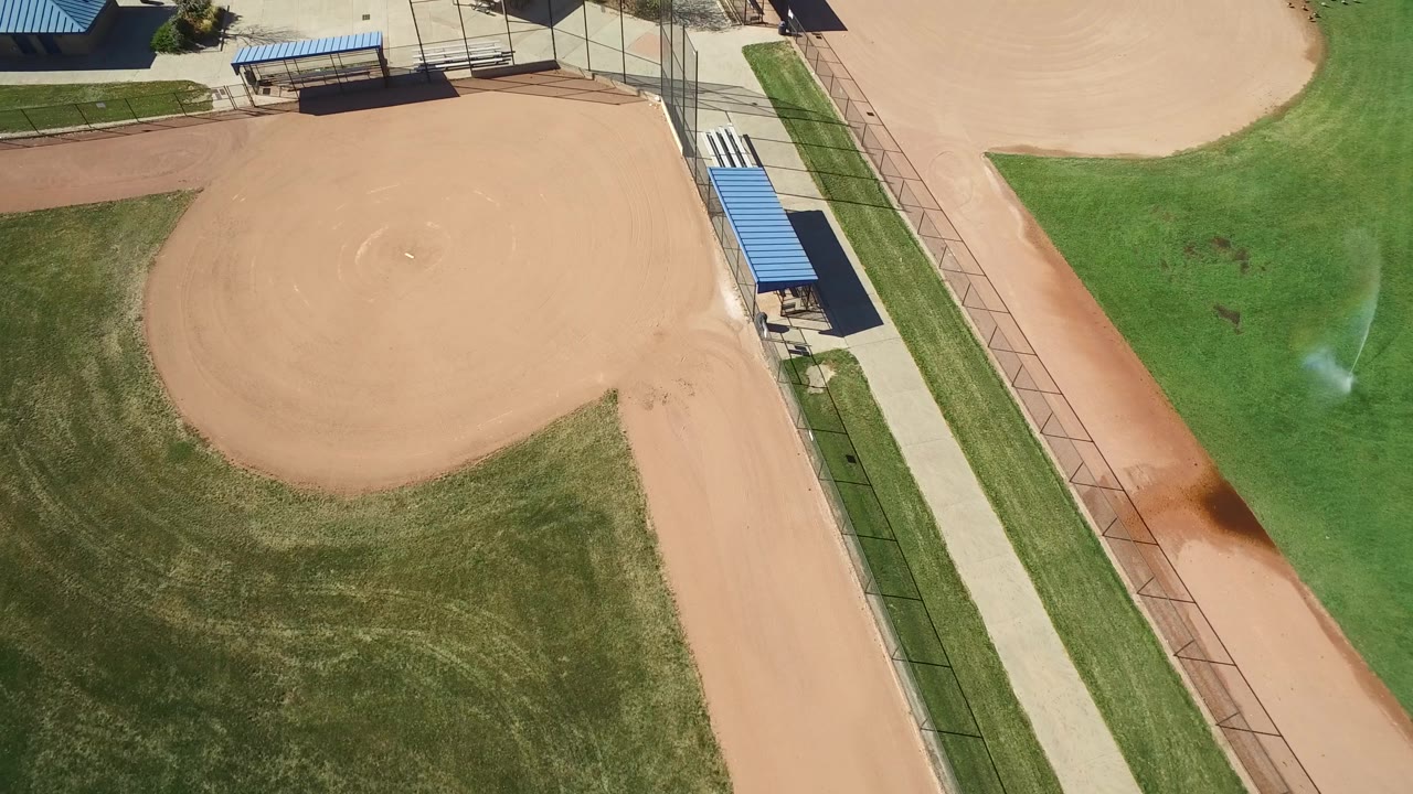 Aerial view in Hidden Lake, Colorado with the baseball infield centered left, a sprinkler spraying on the right-side grass, paved paths, fencing, blue roofs, and no visible people.
