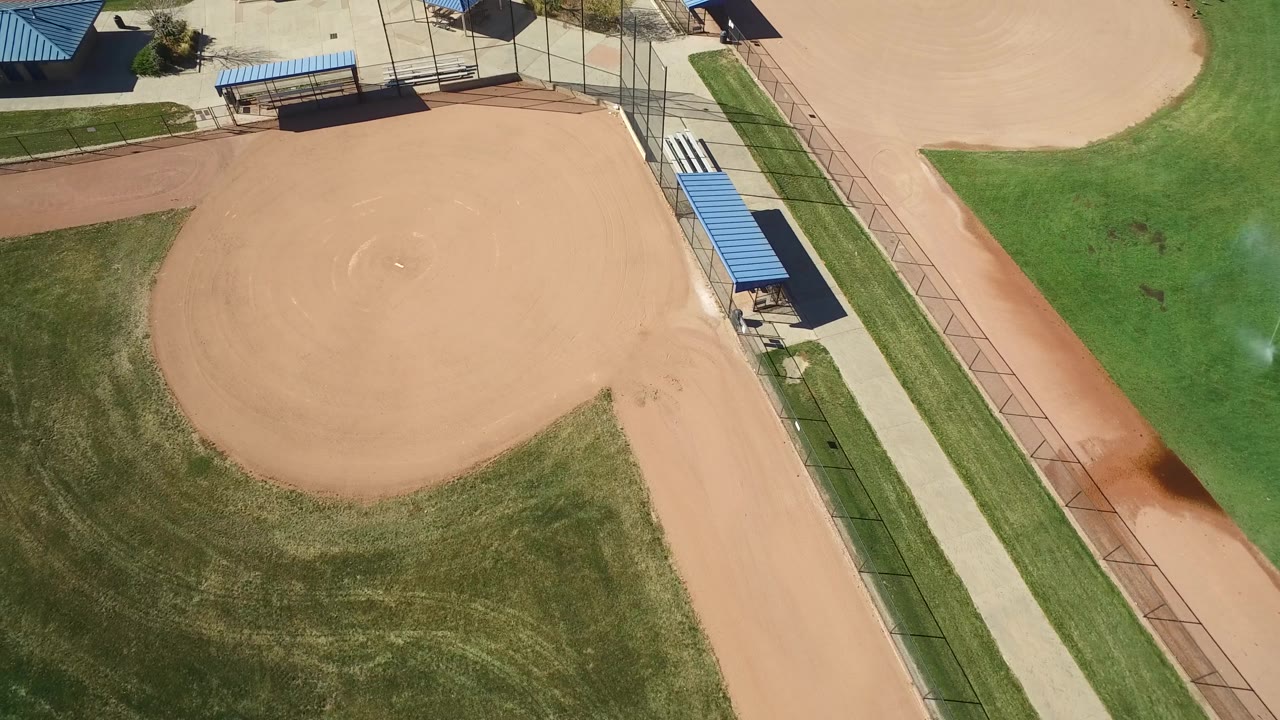 Top-down park view with a tan infield, blue roofs, fencing, and green lawn.