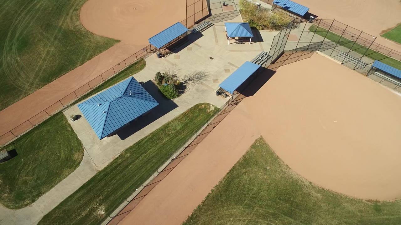 Top-down aerial frame in Hidden Lake, Colorado showing blue-roofed shade structures, a small building, tan ballfield surfaces, concrete paths, fencing, and no visible people.