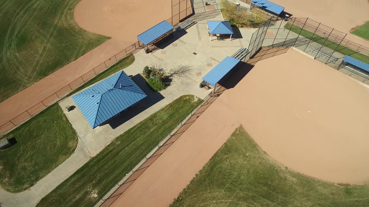 Overhead Hidden Lake, Colorado view with the central blue-roofed building shifted lower in frame, surrounded by mowed grass, tan infields, walkways, fences, and no visible people.