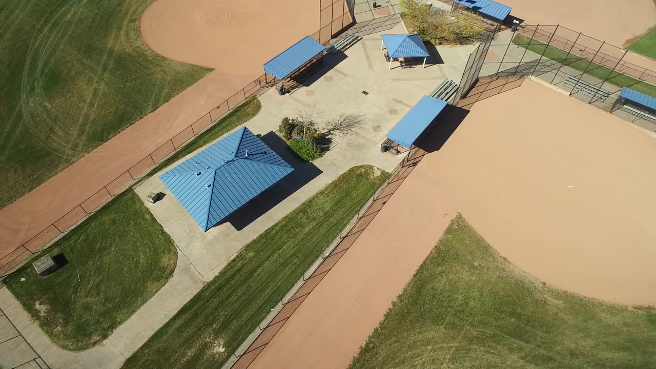Top-down park scene with blue shelter roofs and adjacent ballfield dirt.