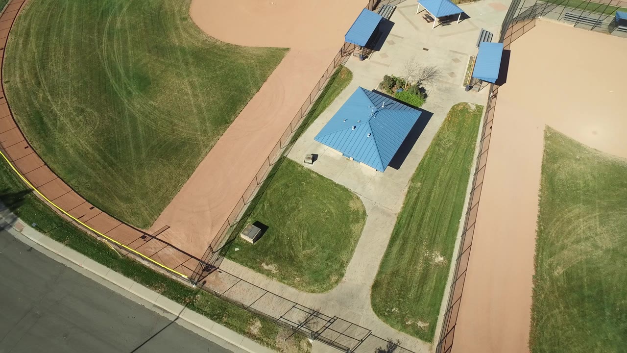 Opening overhead frame in Hidden Lake, Colorado showing a curved green field edge, tan infield surface, blue-roofed utility building, roadside fence, and no visible people.