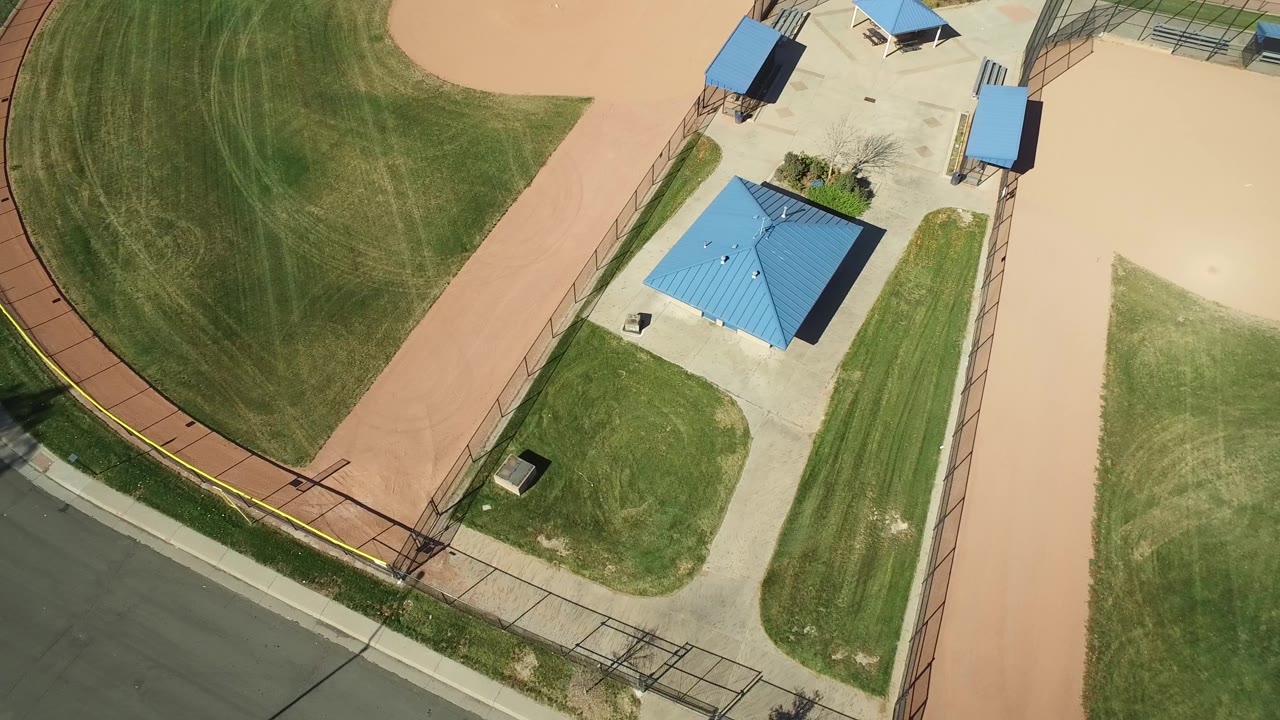 Second overhead frame in Hidden Lake, Colorado with the blue-roofed building closer to center, tan field areas on both sides, fencing along the road, and no visible people.
