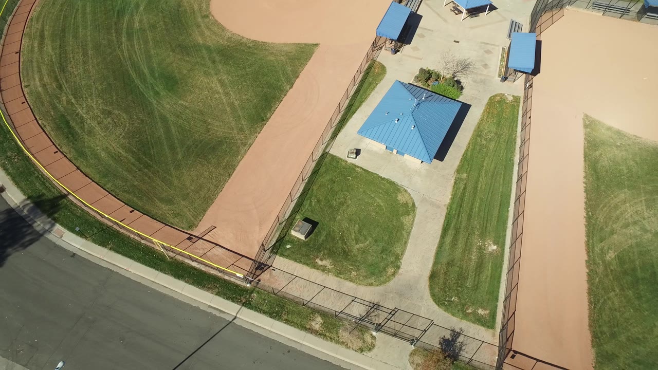 Aerial view of a fenced field, small blue-roofed building, tan paths, and empty road.