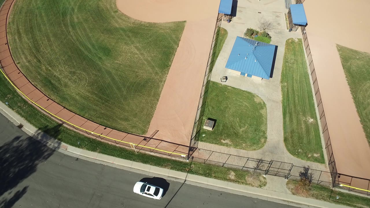 Overhead frame from Hidden Lake, Colorado shows the curved green outfield, tan infield dirt, a roadside fence, a blue-roofed shelter, and a white car at the bottom, with no visible people.