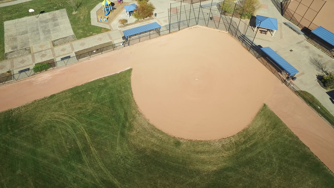 Overhead aerial frame of a tan baseball infield, green outfield grass, paved park paths, blue-roofed shade structures, and a playground in Hidden Lake, Colorado, with no visible people.