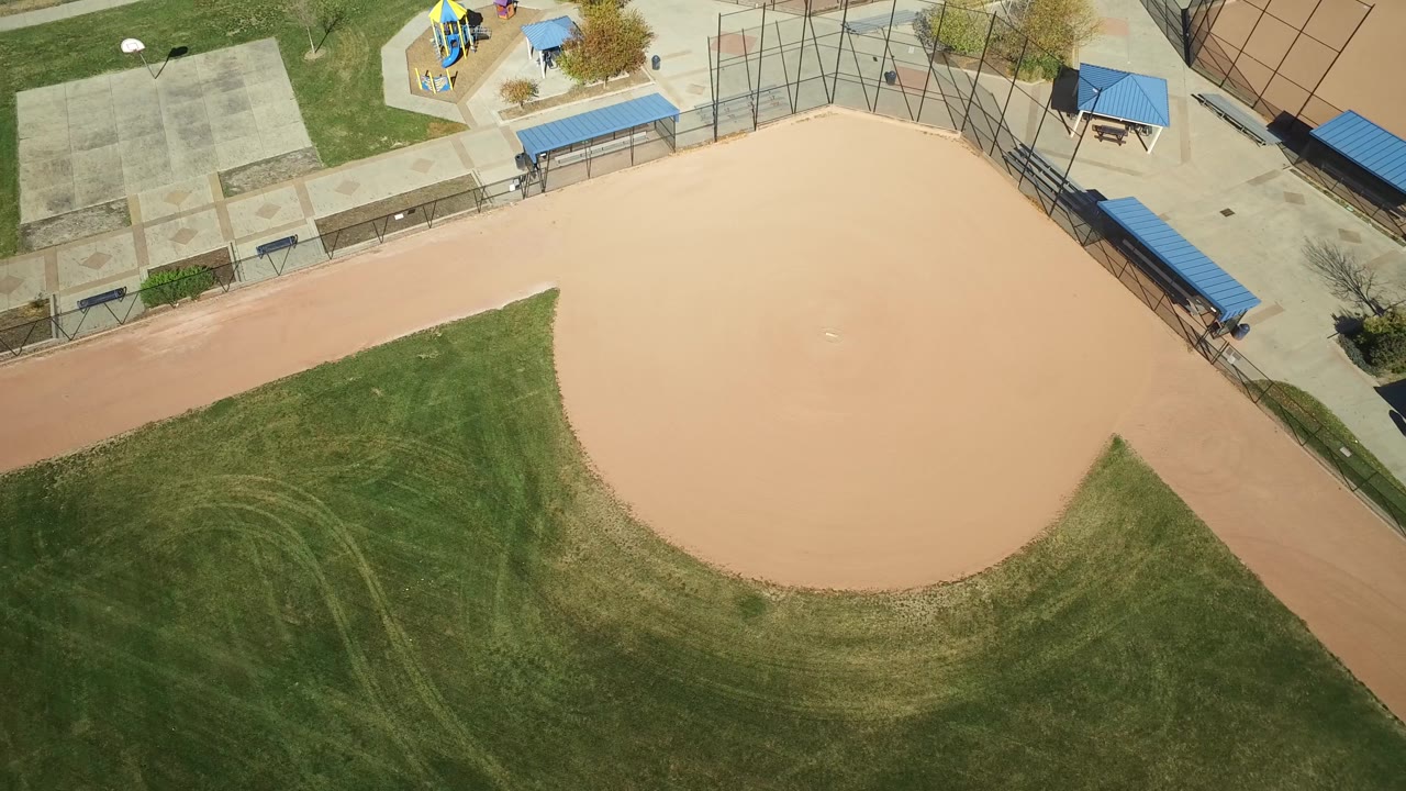 High-angle view over the Hidden Lake baseball infield, with curved grass edges below, concrete courts to the upper left, blue-roofed structures along the fence, and no visible people.