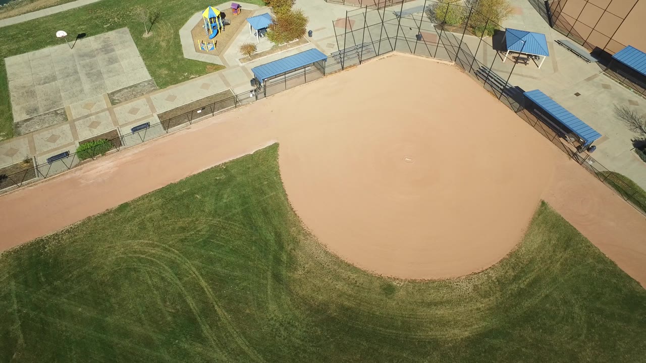 Drone view of a tan infield bordered by grass, fencing, and playground equipment.