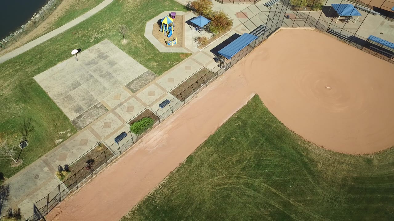 Overhead frame in Hidden Lake, Colorado showing the baseball infield edge on the right, concrete walkways, blue shade roofs, a small playground, grass, lake shore, and no visible people.