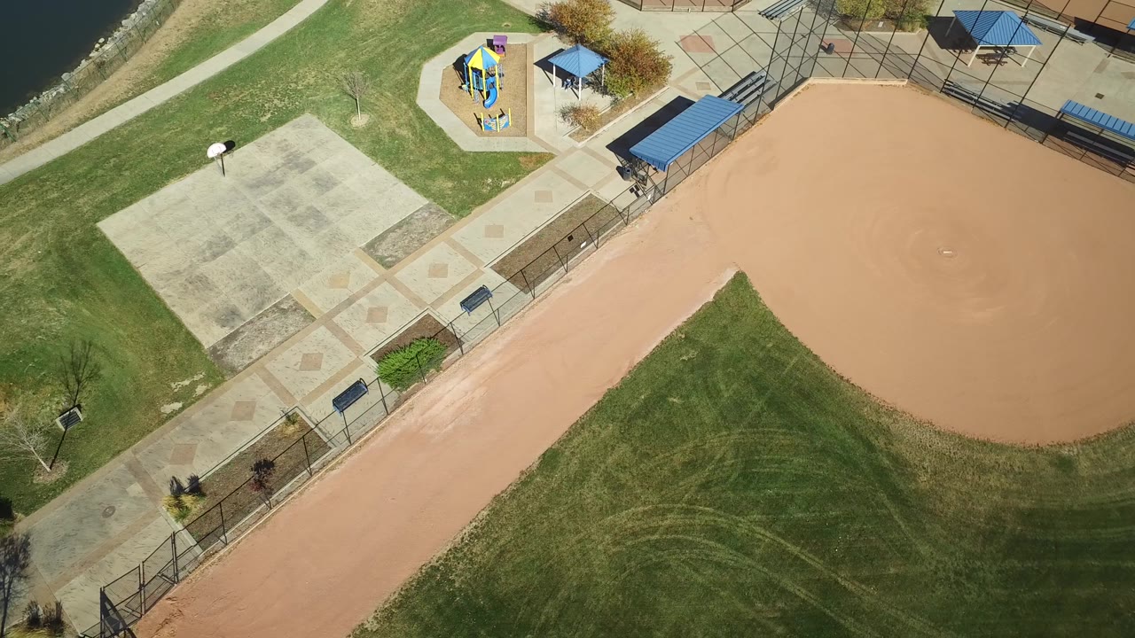 High overhead view in Hidden Lake, Colorado with the tan infield still on the right, lawn in the lower center, patterned concrete paths, playground structures near the top, lake edge at left, and no visible people.