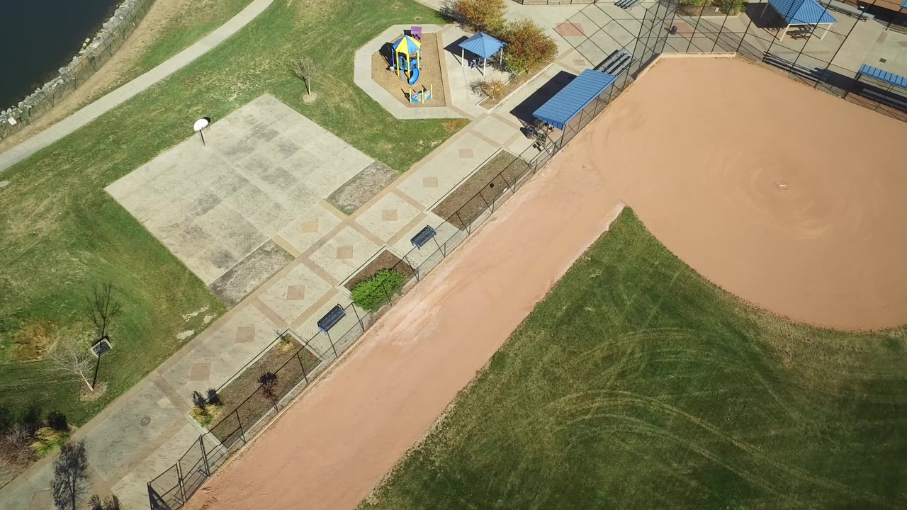Drone view of the baseball infield corner, grass, paved plaza, and playground by water.