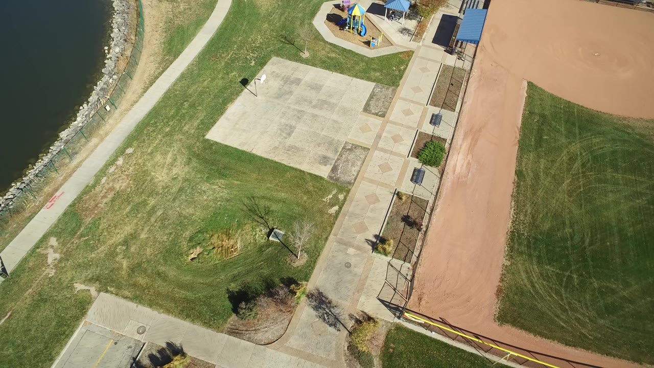 Aerial view in Hidden Lake, Colorado with the shoreline path along the left edge, open lawn and concrete paving in the center, reddish ball field dirt on the right, and no clearly identifiable people.
