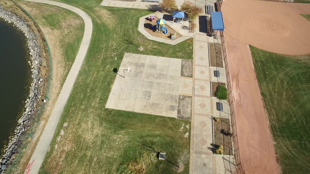 Top-down aerial frame over Hidden Lake, Colorado showing shoreline rocks at left, a curved path, grass, a concrete pad, playground shade roofs, and a reddish ballfield area with no visible people.