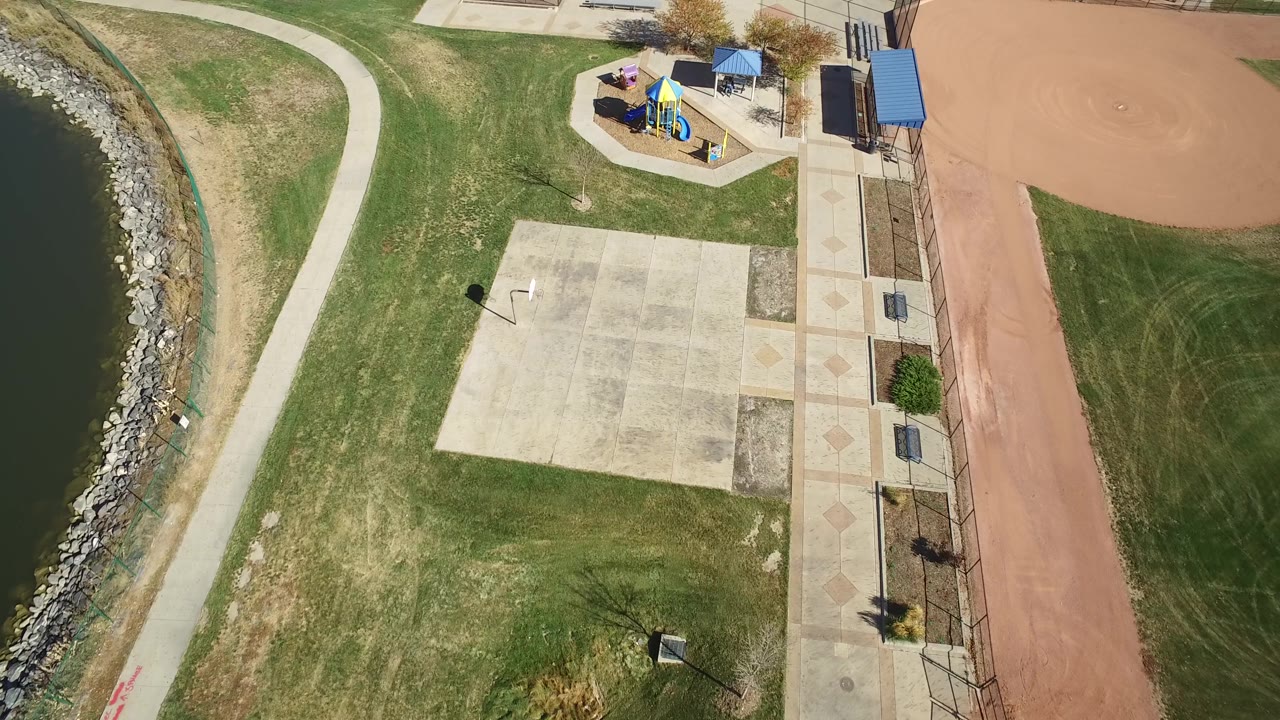 Drone-style overhead view in Hidden Lake, Colorado with water and riprap along the left edge, a pale concrete pad below center, a small playground near the top, and no visible people.