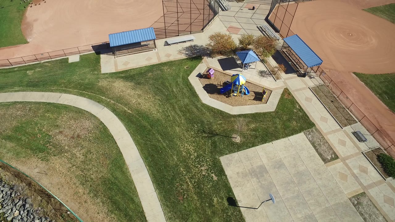 High aerial view over Hidden Lake, Colorado park grounds, with curved paths, patchy grass, blue-roofed shelters, a playground, and a baseball infield edge.