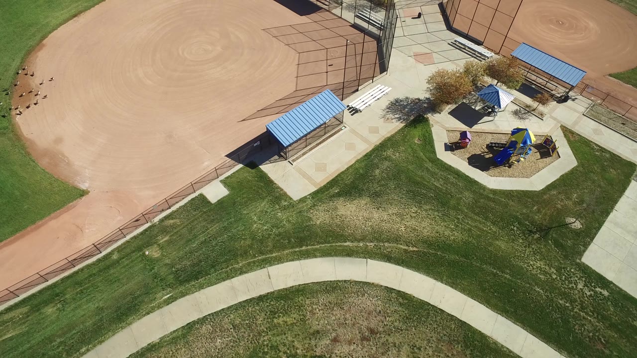 High overhead frame from Hidden Lake, Colorado with the ballfield dirt across the upper left, the playground and shade canopies to the right, and a broad band of grass and path below, with no clearly identifiable people.