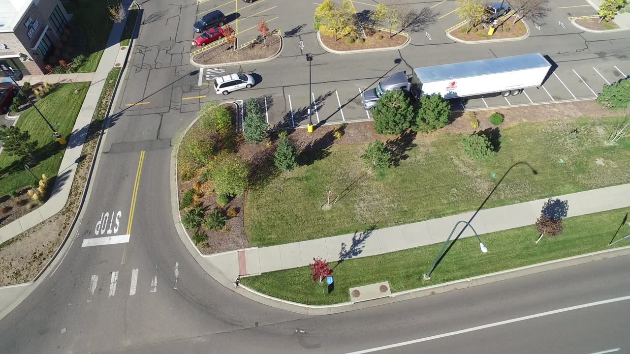 Aerial view of a road corner, sidewalk, grass, parking lot, cars, and a white trailer.