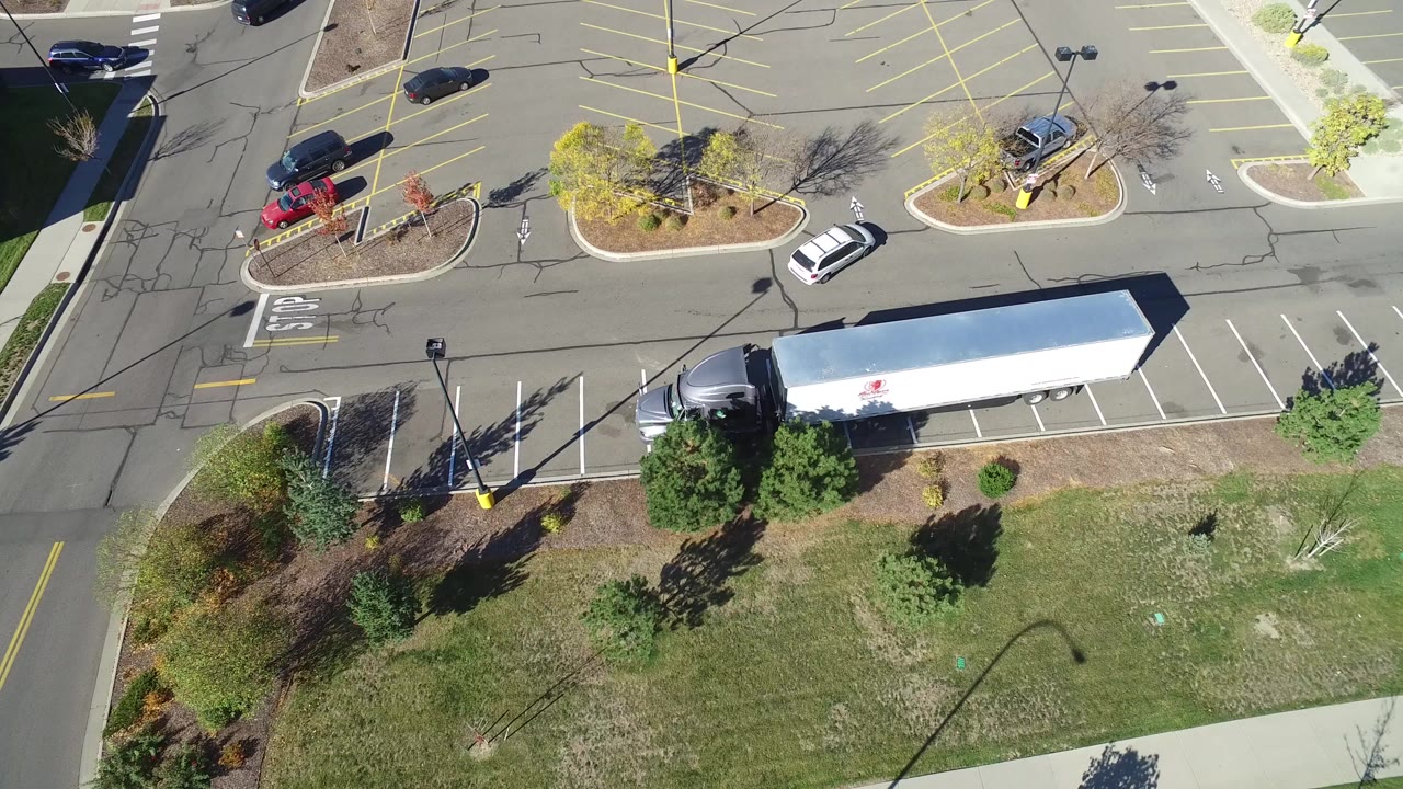 Top-down view of a Hidden Lake, Colorado parking lot at the start of the clip, with a white trailer along the lower edge, a few cars, planted islands, long shadows, and no visible people.