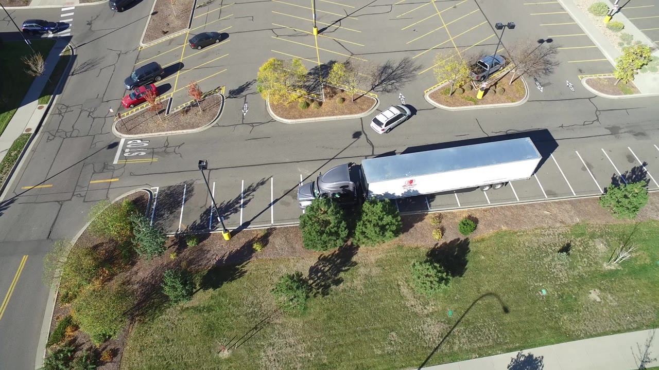 High aerial frame over Hidden Lake, Colorado parking stalls, with the long white trailer near the bottom, a red vehicle near a planted island, scattered cars, lamp shadows, and no visible people.