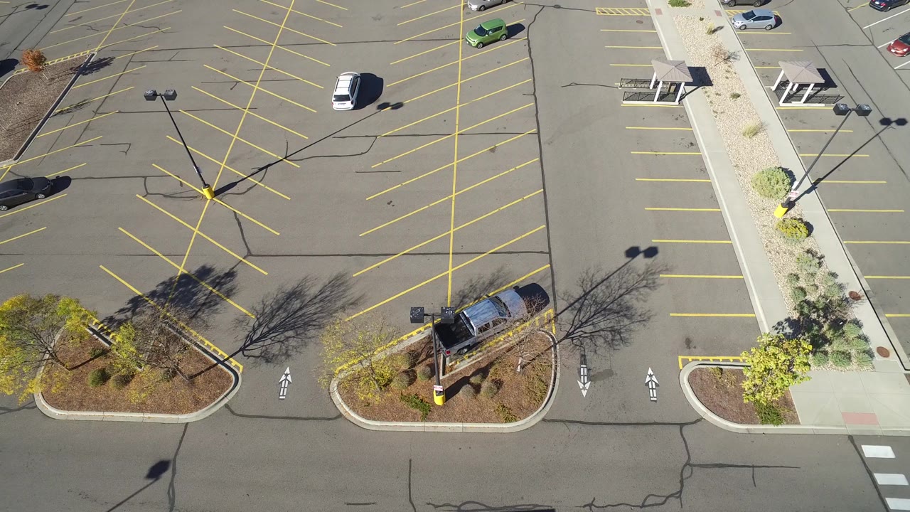 High overhead frame in Hidden Lake, Colorado with diagonal yellow parking rows, a white vehicle near the upper lot, landscaped curb islands, strong shadows, and no visible people.