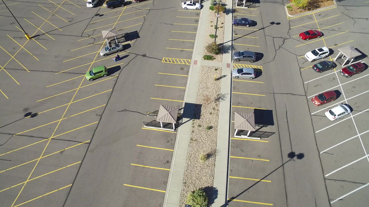 High aerial frame over a Hidden Lake, Colorado parking lot, showing yellow stall lines, a central landscaped walkway, a few parked cars, and no visible people.