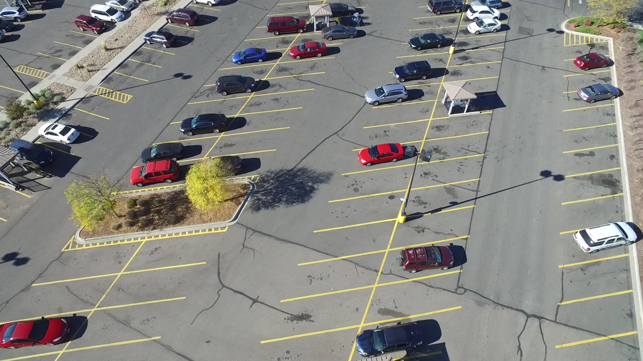 Aerial frame over Hidden Lake, Colorado parking rows, with a red car near the center, a blue car near the upper row, empty marked stalls, hard sun shadows, and no visible people.