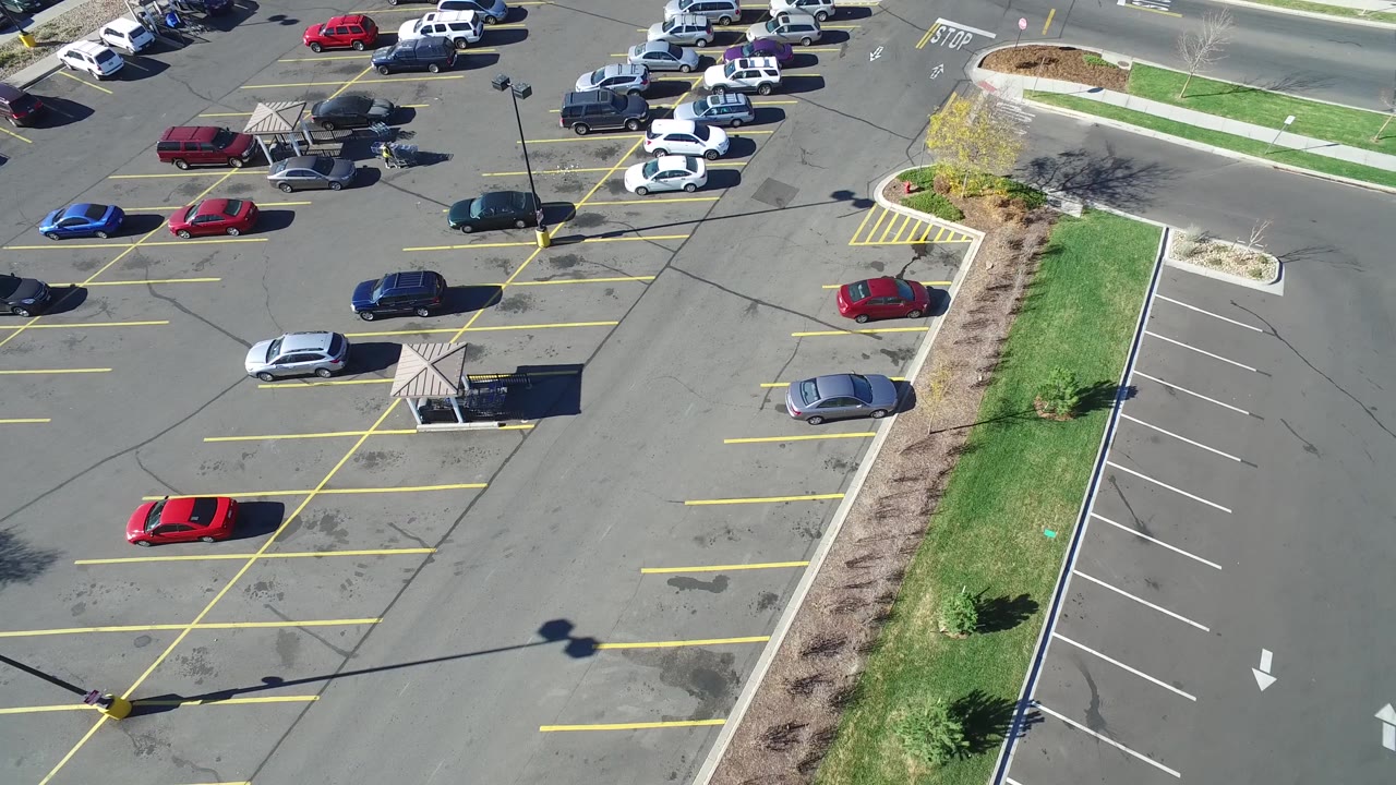 Aerial frame over the Hidden Lake, Colorado lot showing a red car near mostly open parking spaces, several white cars near the road, landscaped beds, and no visible people.