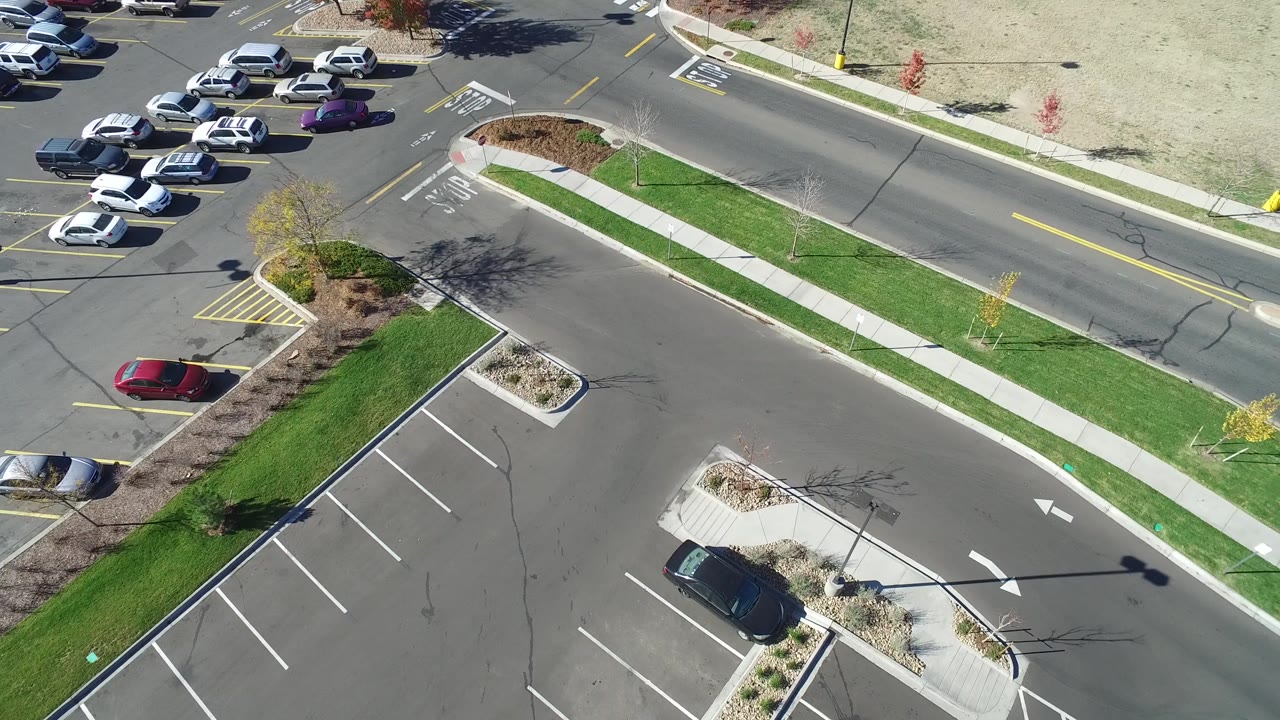 Aerial road scene with parked cars, sidewalk, grass strip, and pale open ground.