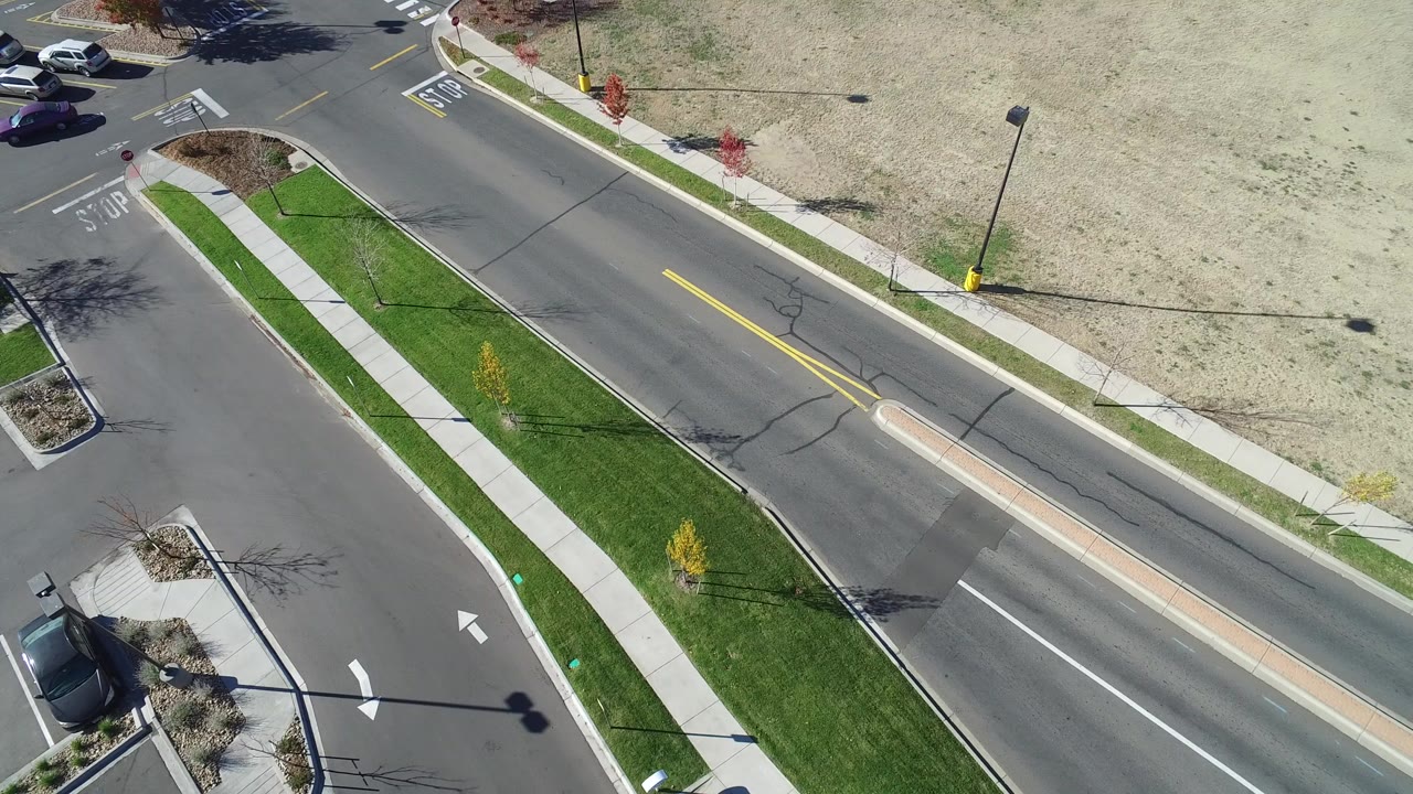 High oblique aerial frame over Hidden Lake, Colorado showing a paved residential road, green grass strip, sidewalks, lamp posts, parked cars near the corner, and no visible people.