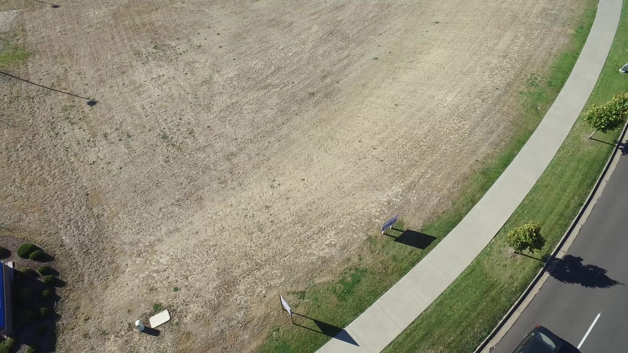 Hidden Lake, Colorado aerial frame with the sidewalk cutting between the dry field and road while a partial car appears near the lower right edge and no visible people are present.