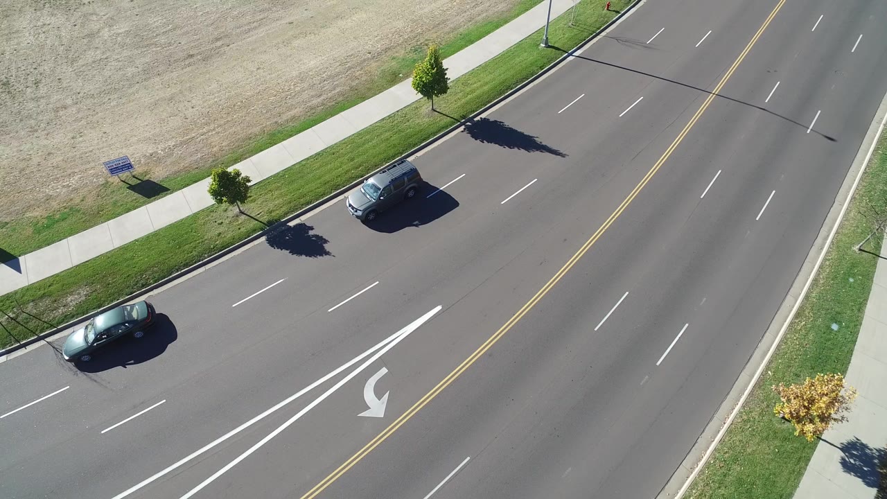 Overhead frame from Hidden Lake, Colorado shows two dark cars on a wide paved road beside a sidewalk, grass strip, small trees, and dry open ground, with no visible people.