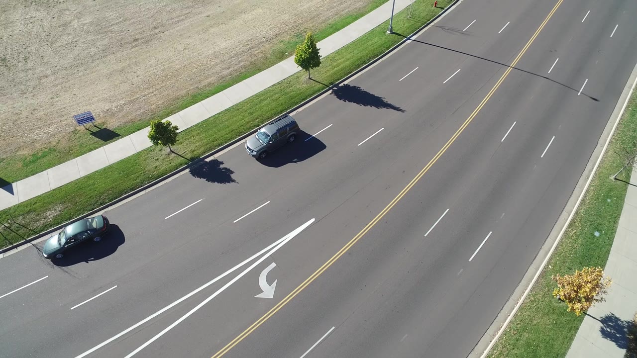Aerial frame from Hidden Lake, Colorado shows two cars farther apart on the curved road, with the sidewalk, grass verge, young trees, and dry field visible along the upper edge, with no visible people.