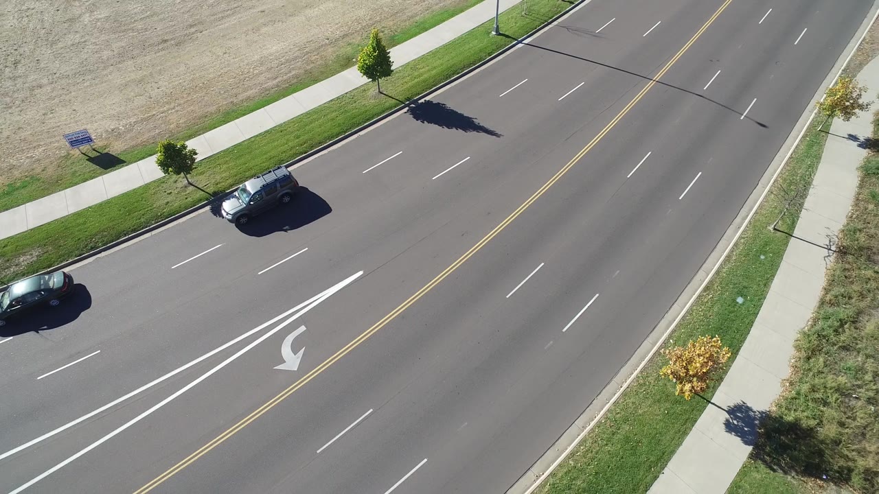 Top-down road frame with two dark cars, double yellow lines, and a landscaped sidewalk edge.