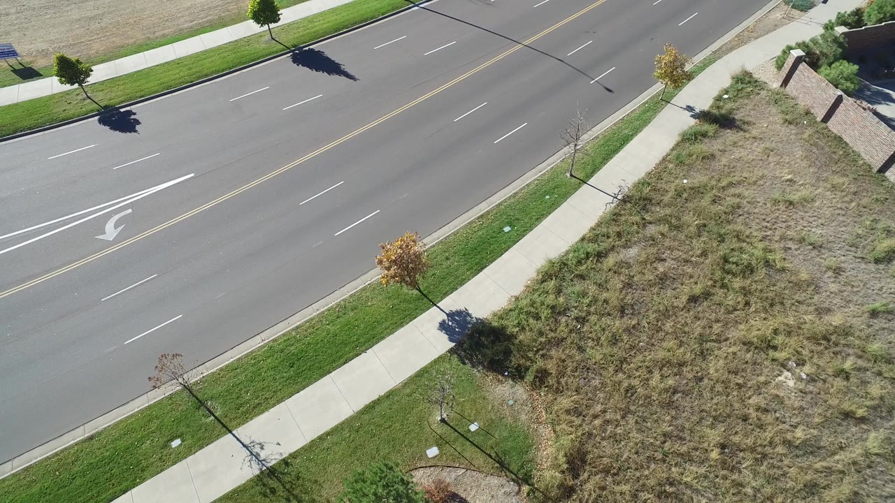 High aerial frame in Hidden Lake, Colorado showing an empty multi-lane road, sidewalks, grass strips, small trees, and a dry vacant lot with no visible people.