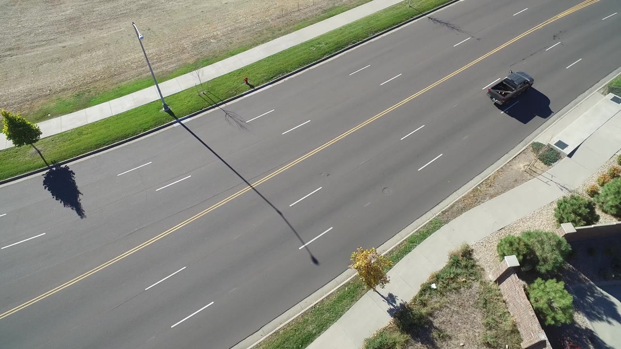 Aerial Hidden Lake, Colorado frame with the dark vehicle farther along the upper roadway, diagonal lane markings, sidewalk strips, dry grass, and a small pedestrian visible near the roadside.