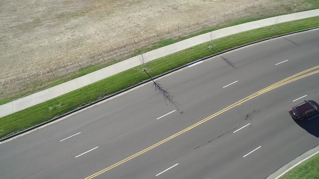 Aerial frame from Hidden Lake, Colorado showing the diagonal roadway, yellow double lines, white lane marks, sidewalk, dry grass beyond it, a dark car at the right side, and no visible people.