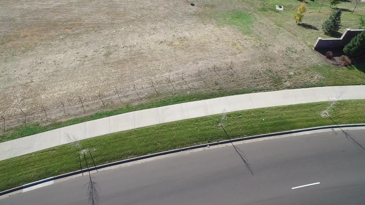 Aerial frame over Hidden Lake, Colorado showing a curved paved road, pale sidewalk, tan open grass, a green lawn strip, and no visible people.