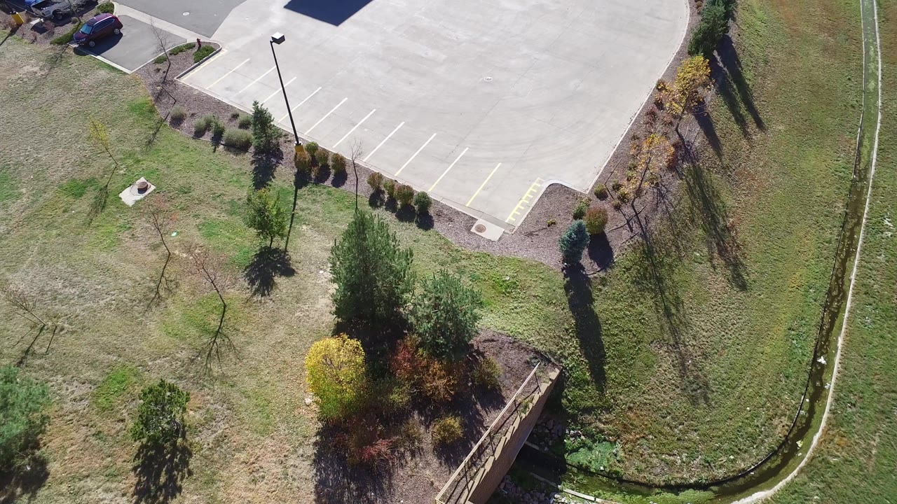 Overhead aerial frame in Hidden Lake, Colorado showing the corner of an empty parking lot, grass, landscaped shrubs, trees, a drainage ditch, and no visible people.