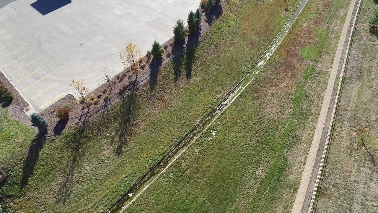 Steep aerial view in Hidden Lake, Colorado showing grass, a narrow drainage channel, parking-lot pavement, trees with long shadows, and no visible people.