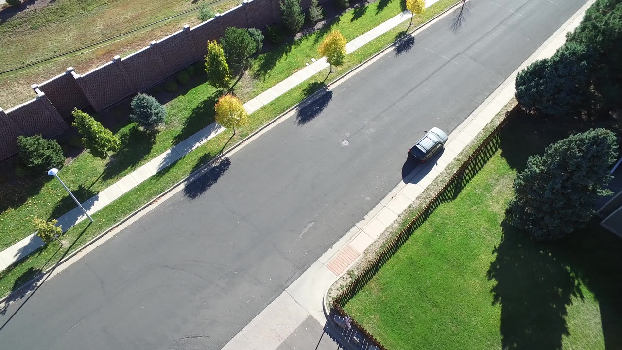 Overhead suburban road with lawns, sidewalks, trees, and a dark car.