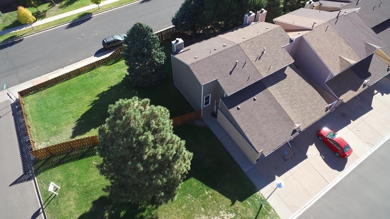 Overhead frame in Hidden Lake, Colorado showing adjacent homes, a fenced green yard, a large tree casting shade, a residential road, and a red car on the driveway, with no clearly visible people.
