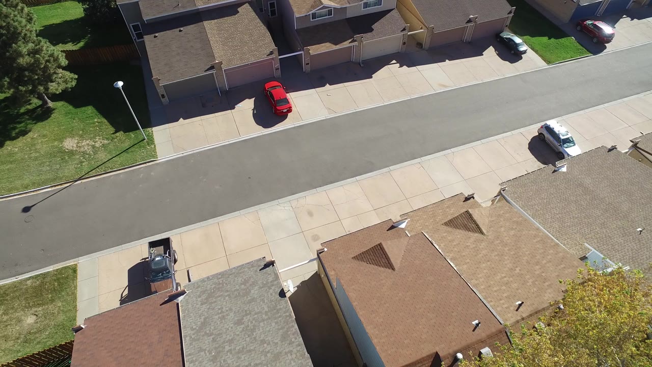 Overhead drone view in Hidden Lake, Colorado showing suburban roofs at the bottom, a quiet street through the center, sidewalks, lawns, parked cars, and no visible people.