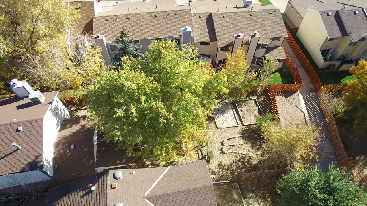 Aerial frame in Hidden Lake, Colorado with townhouse roofs, fenced backyards, bare garden plots, green lawn strips, mature trees, and no visible people.